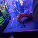 Gros plan de mains tapant sur un ordinateur portable affichant des graphiques de cybersécurité, éclairés par une lumière violette.